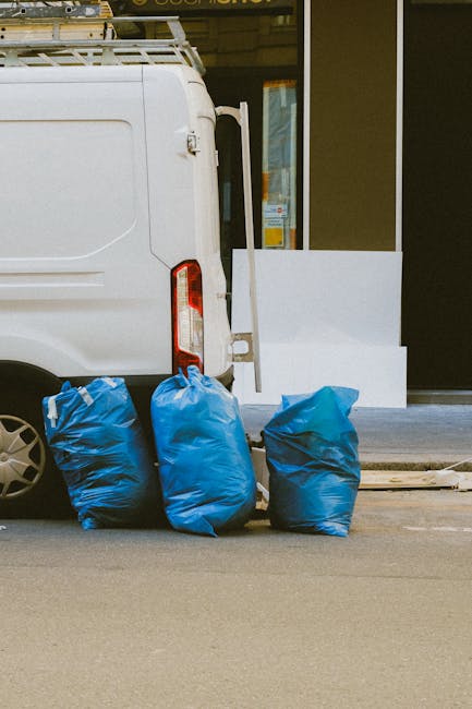 Two waste collection workers in dark green uniforms with reflective orange stripes are seen in an urban setting during dusk, standing beside a large commercial rubbish truck. One worker, on the left,