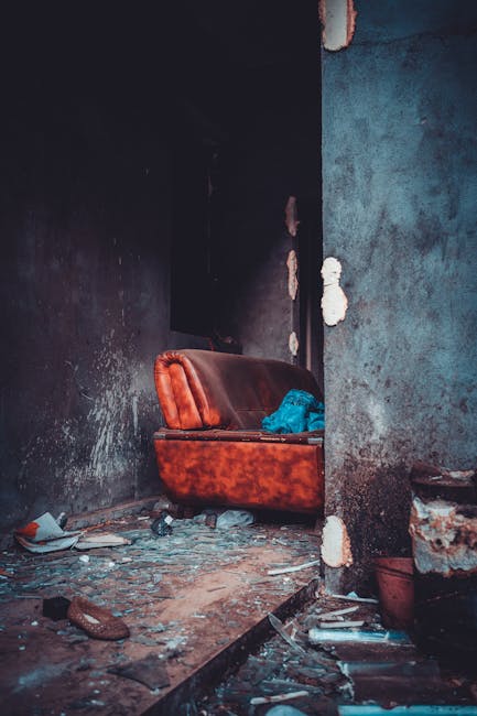 In a neglected, possibly abandoned interior space, a worn, brown leather armchair with visible stitching and slightly sagging cushions is positioned against a corner formed by a blackened, dirt-staine