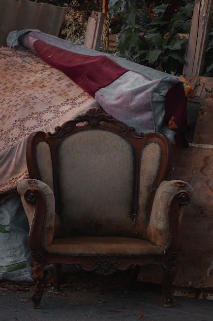 In a neglected, possibly abandoned interior space, a worn, brown leather armchair with visible stitching and slightly sagging cushions is positioned against a corner formed by a blackened, dirt-staine