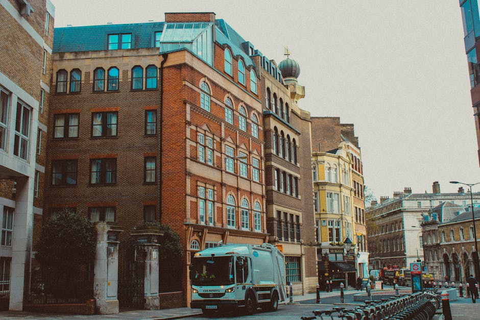 A narrow urban street scene in daylight shows a sidewalk on the left lined with Victorian-style brick terraced houses featuring arched windows and black wrought iron railings. A black lamppost stands 
